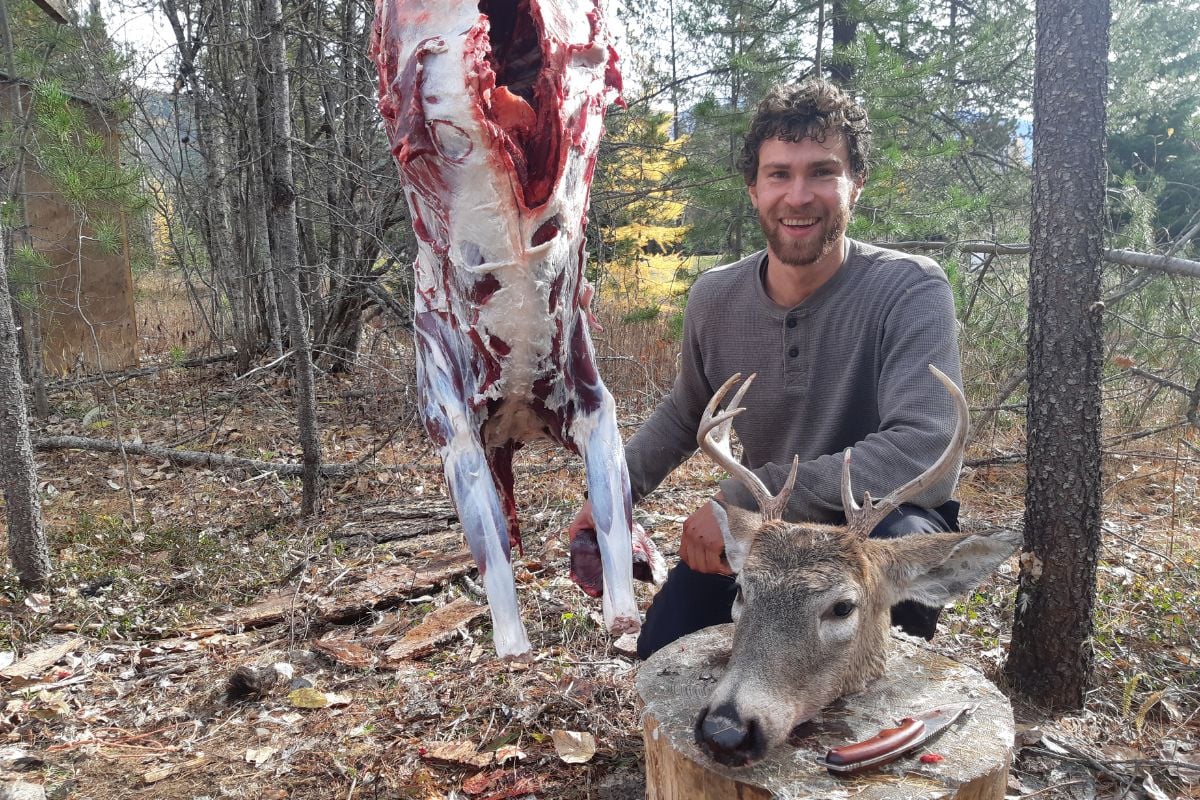 A man with a deer hanging in a tree and a buck's head on the ground nearby.