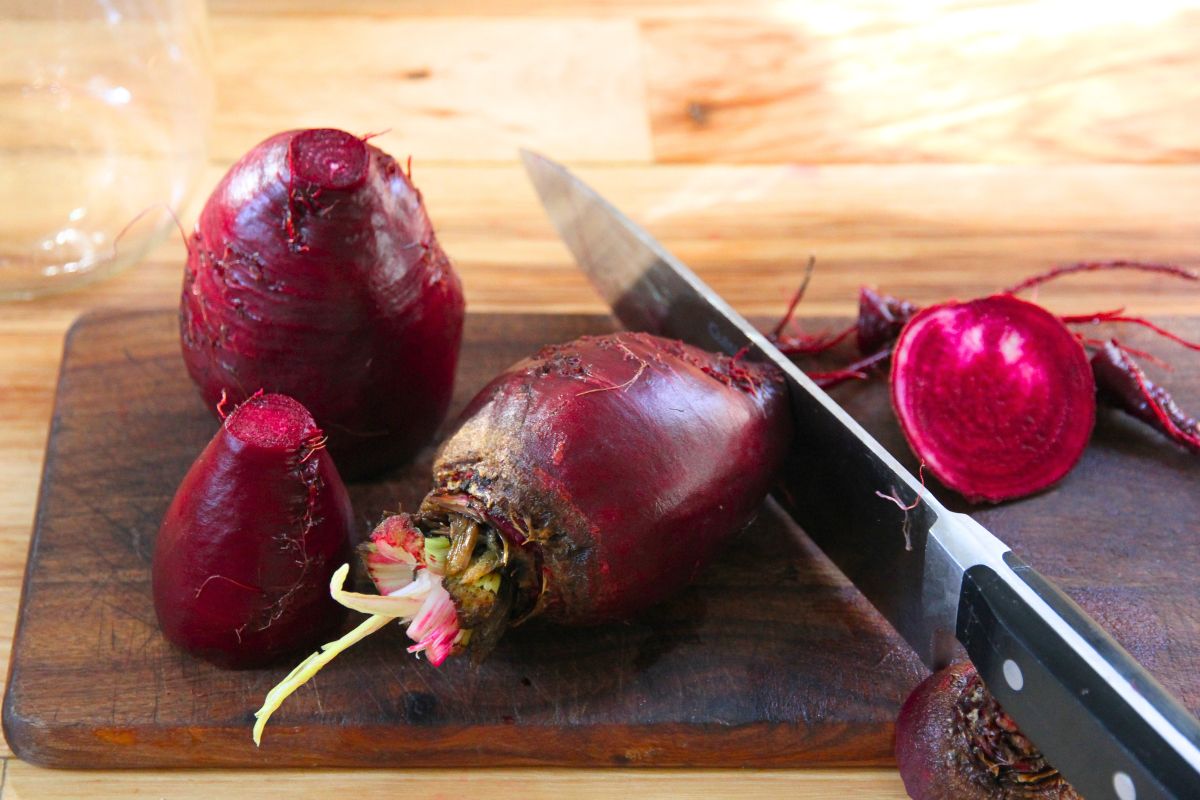 A large organic beet on a wood cutting board, with the tail being sliced off by a large kitchen knife. 
