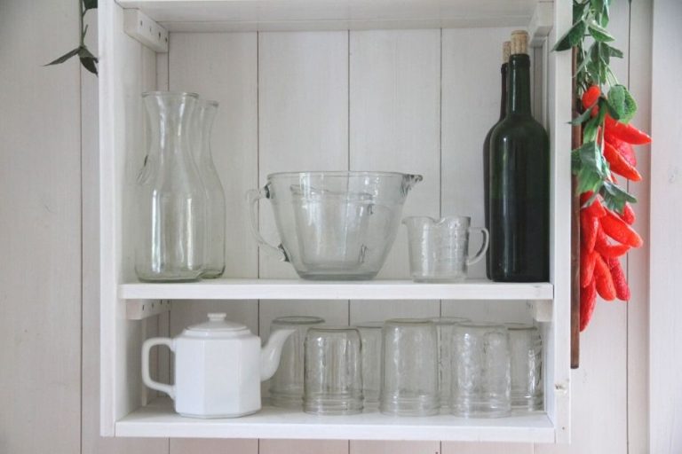 An open upper kitchen shelf displaying glass jars, old milk jugs, a white teapot and glass measuring bowls
