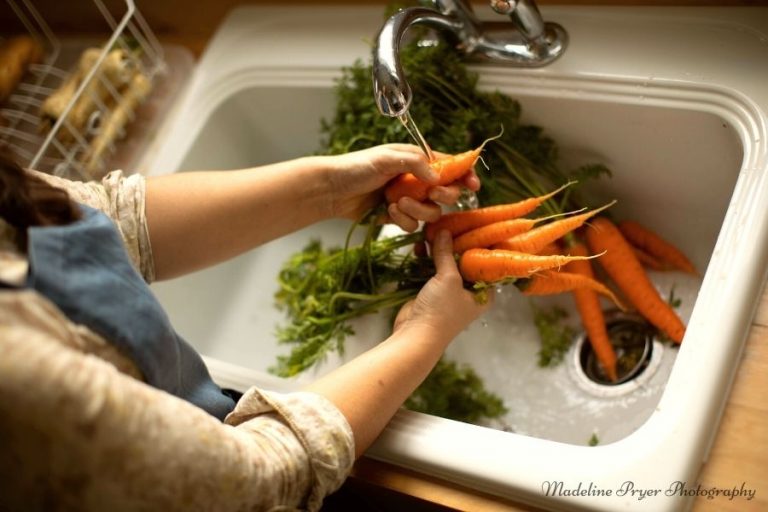 A woman washing home grown carrots at a white kitchen sink