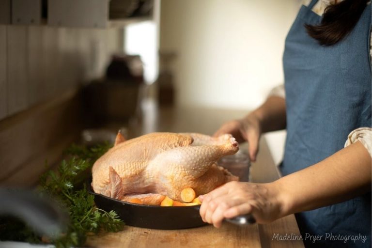 a woman at the kitchen counter in a blue apron, preparing a whole chicken for baking