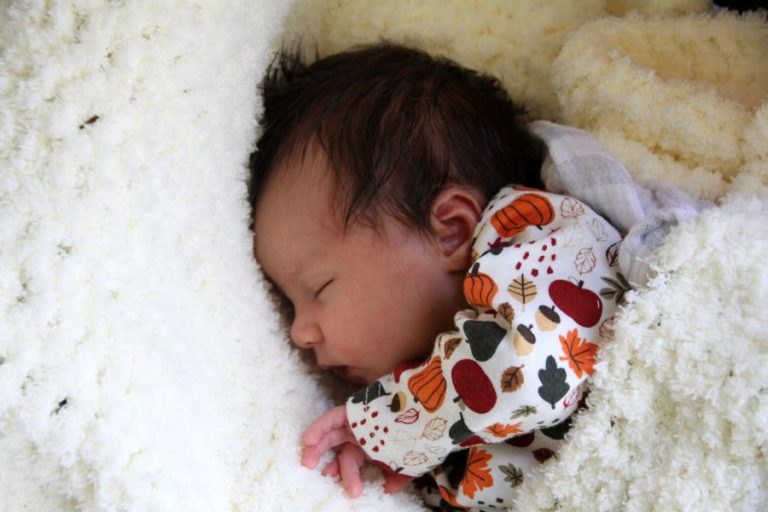 a baby girl with a dark head of hair sleeping on a white knit blanket