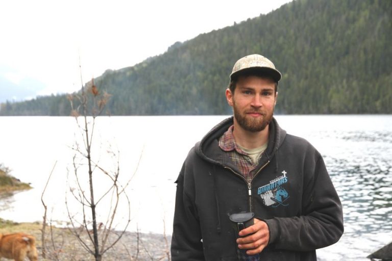 a man by a lake wearing a camouflage cap while holding a thermos of hot coffee