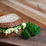 an egg salad sandwich sitting on a wooden cutting board with green parsley on the side