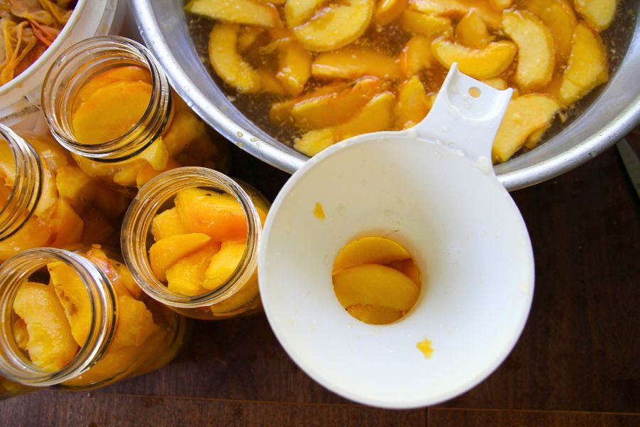Slices peaches being added to a glass jar using a white kitchen funnel.