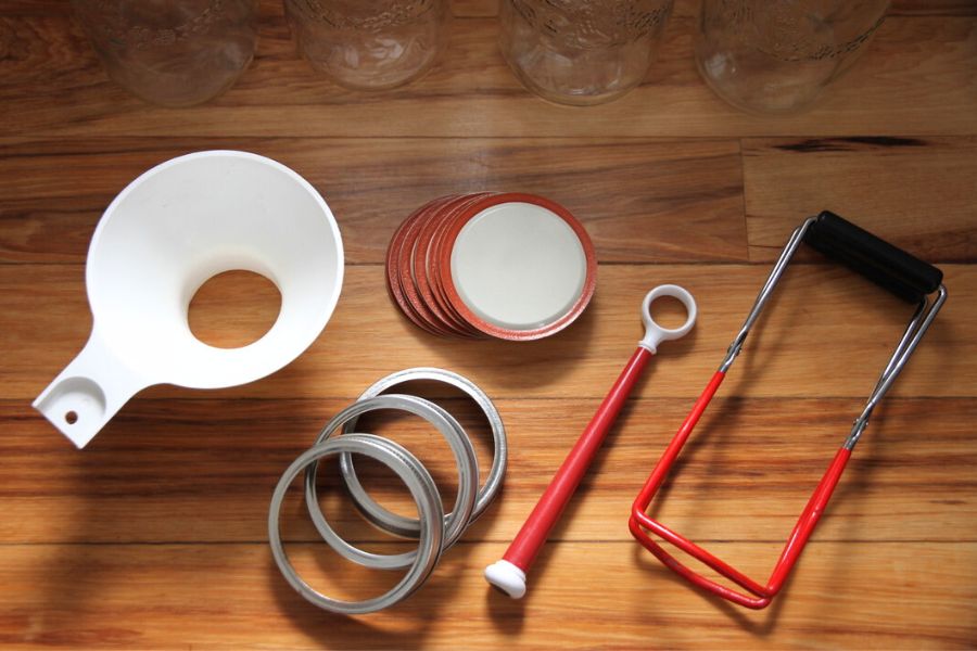 Canning funnel, jar lifter, lid lifter, plus canning lids and metal screw bands laid out on the kitchen counter for canning.