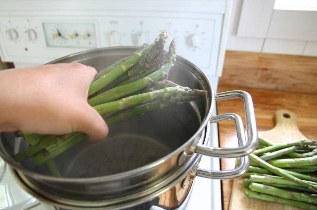 Dropping a handful of prepared asparagus into boiling water on the stove top.