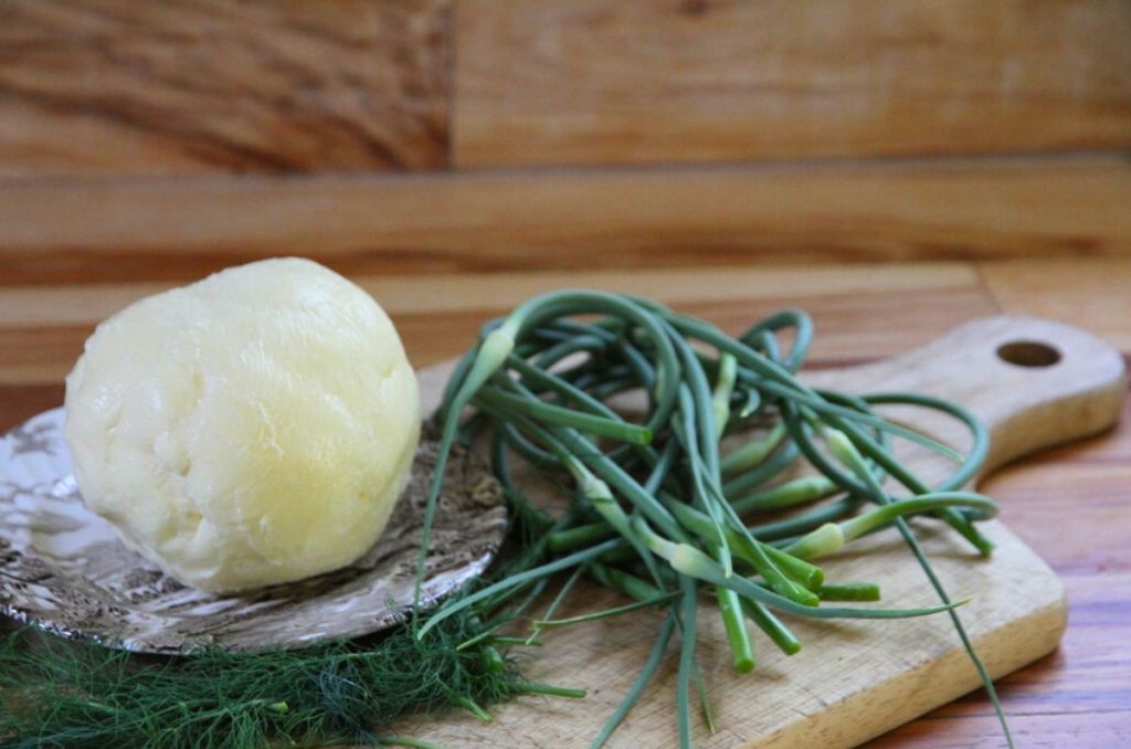 A pile of curly garlic scapes, fresh herbs and a round ball of butter sitting on a wooden cutting board.