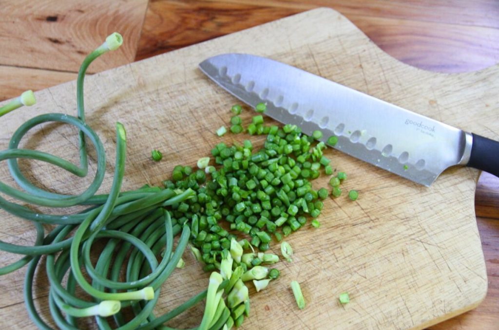 A pile of fresh garlic scapes being minced on a wooden cutting board