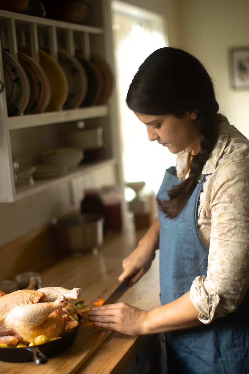 A woman in a blue apron, preparing a whole chicken in a cast iron skillet.