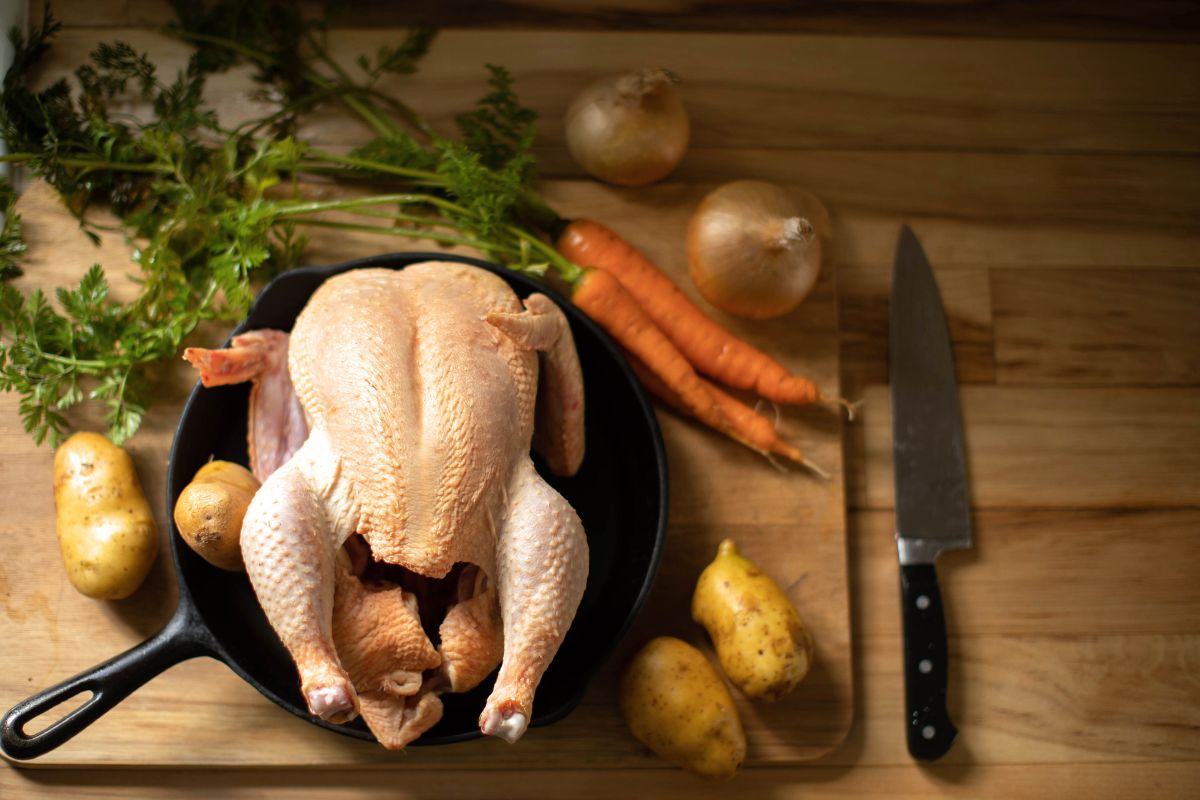 A whole chicken in a cast iron skillet, surrounded by vegetables.