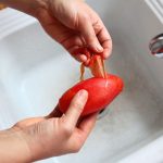 Peeling a frozen, red paste tomato at a white kitchen sink under hot water.