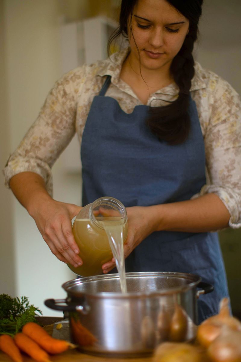 A woman in a dark blue apron pouring chicken broth into a large pot for soup.
