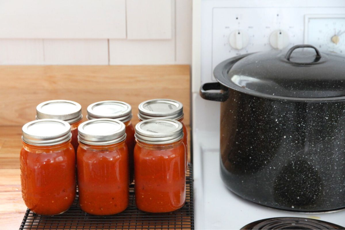 A black enamel water bath canning sitting on a white stove, with 6 pint jars of canned tomato sauce sitting beside on the kitchen counter.