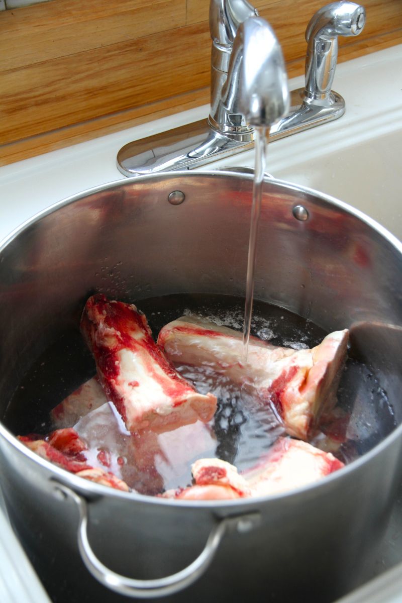 A pile of large beef bones in a stock pot at the kitchen sink, with water being added for broth.