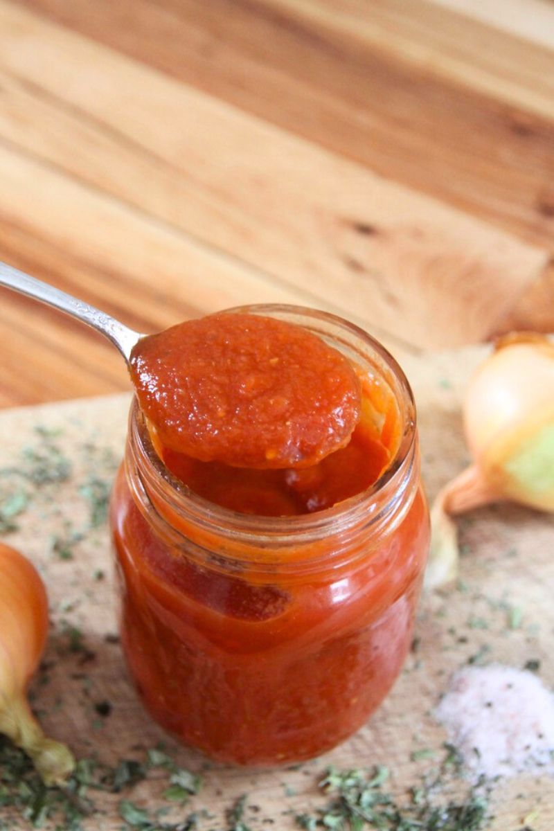 An open jar of homemade red tomato sauce, being lifted with a spoon.