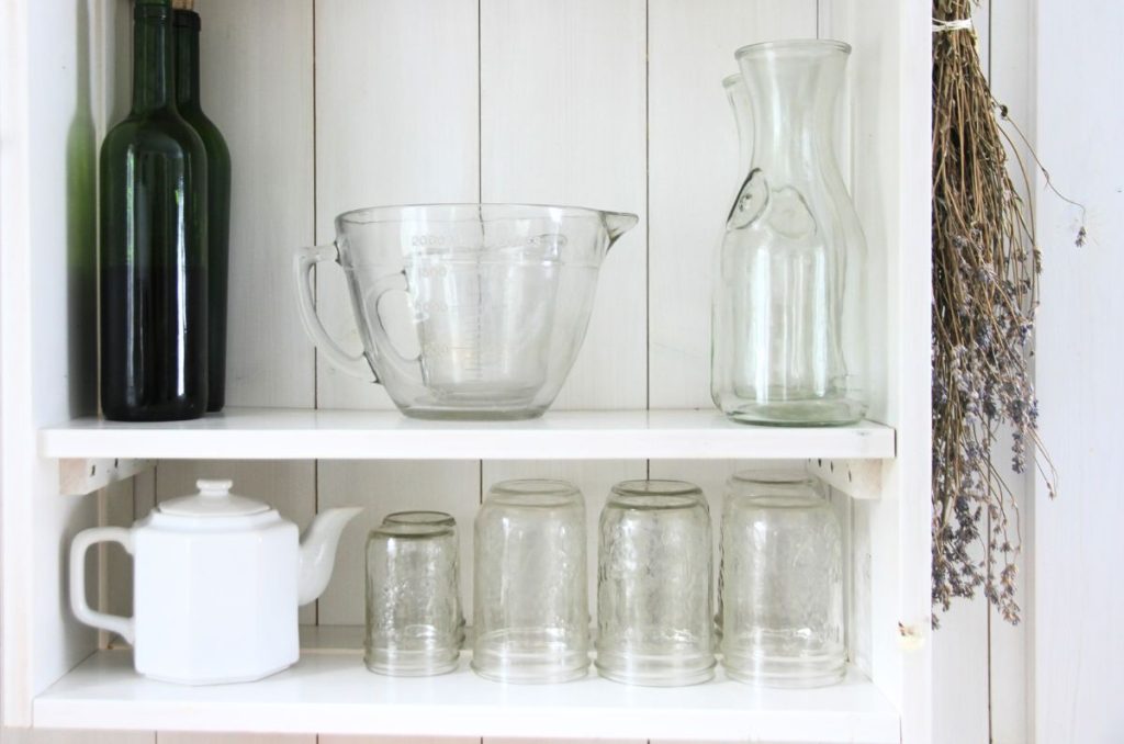 A white kitchen shelf without doors with clear drinking glasses, water jugs, a clear mixing bowl and white teapot on display.