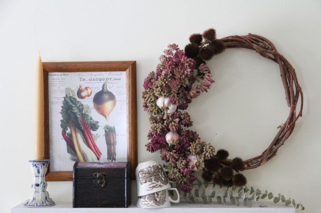A white wall shelf in the kitchen, with vegetable art, a dried herb wreath, brown transferware tea cups, a small brown box and a tapered beeswax candle