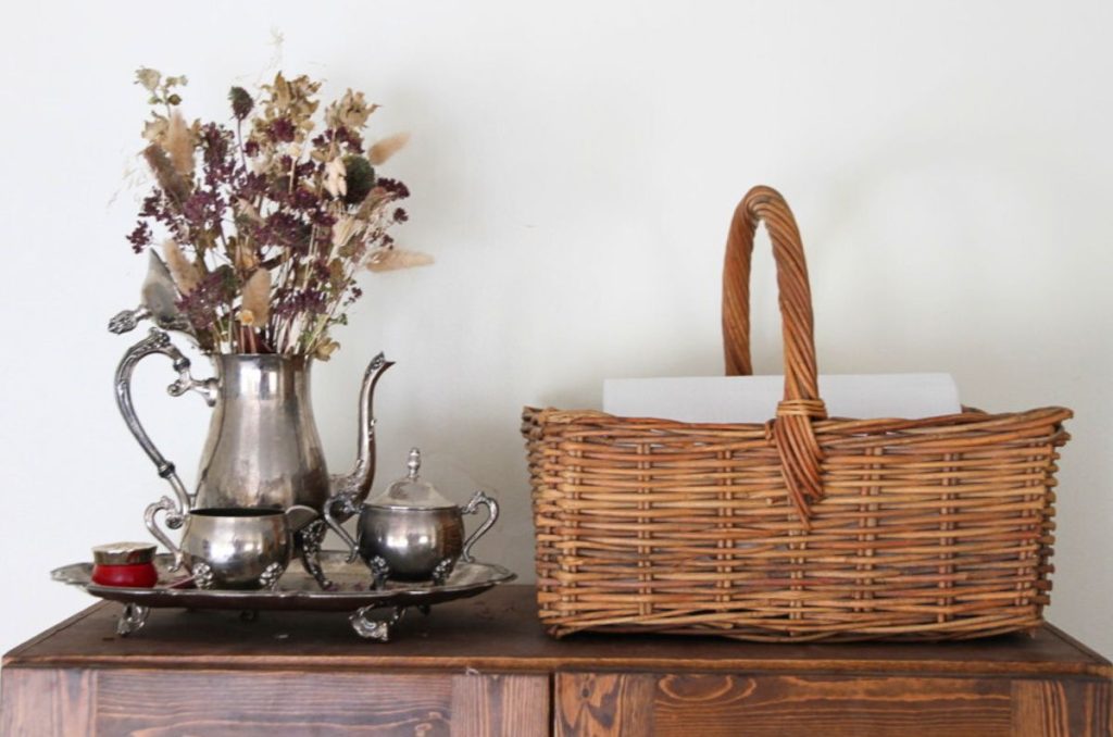 A square woven baskets with a handle sitting on top of a kitchen hutch, next to a silver teapot filled with dried flowers.