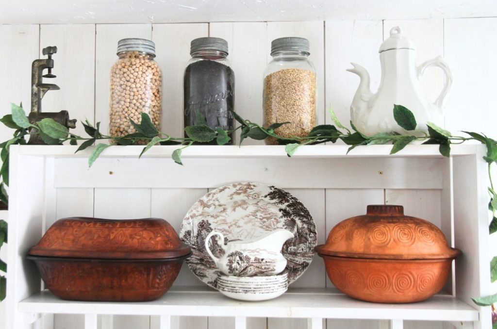 An open kitchen shelf with clay bakeware, a platter, gravy boat and above this shelf, jars of dry goods.