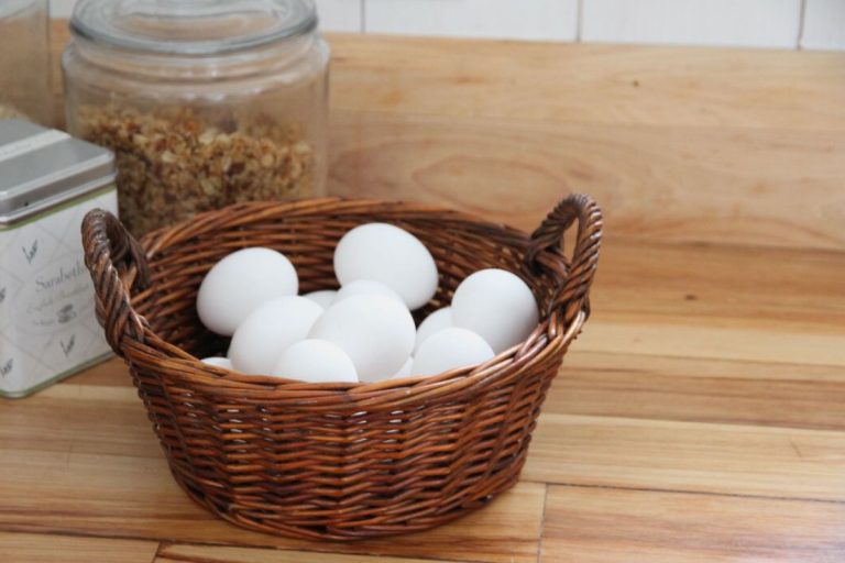 A round woven baskets of white eggs sitting on a wooden kitchen counter.