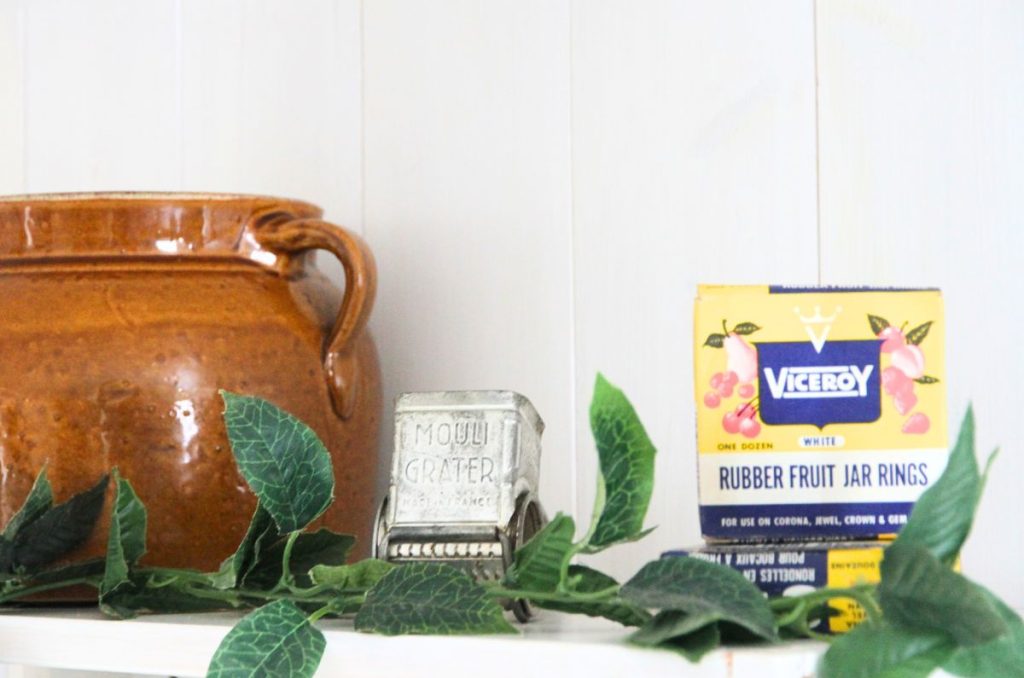 Displaying a brown bean pot, a french cheese grated and a yellow box of canning seals on top of a kitchen shelf. 