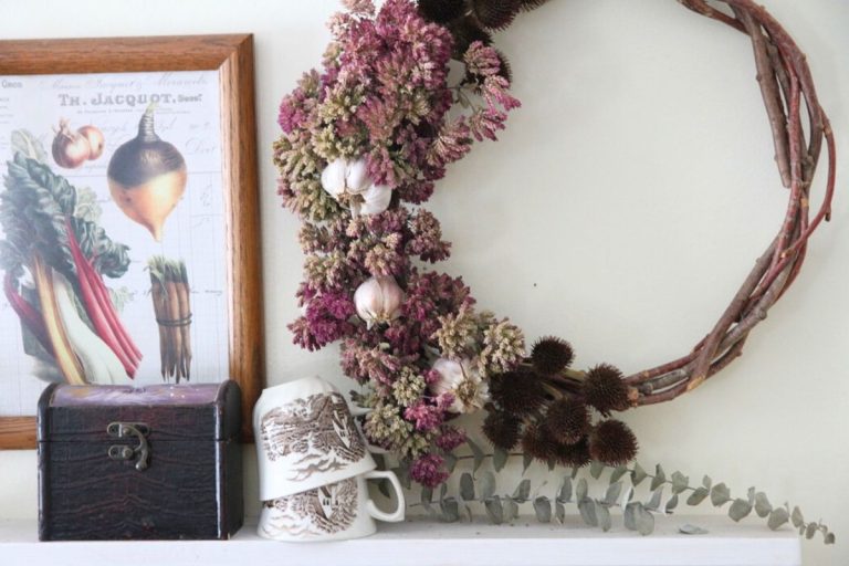 A white kitchen wall shelf displaying brown and white transferware tea cups, a herb wreath of dried oregano flowers and garlic and a framed vintage vegetable print in a wood frame.