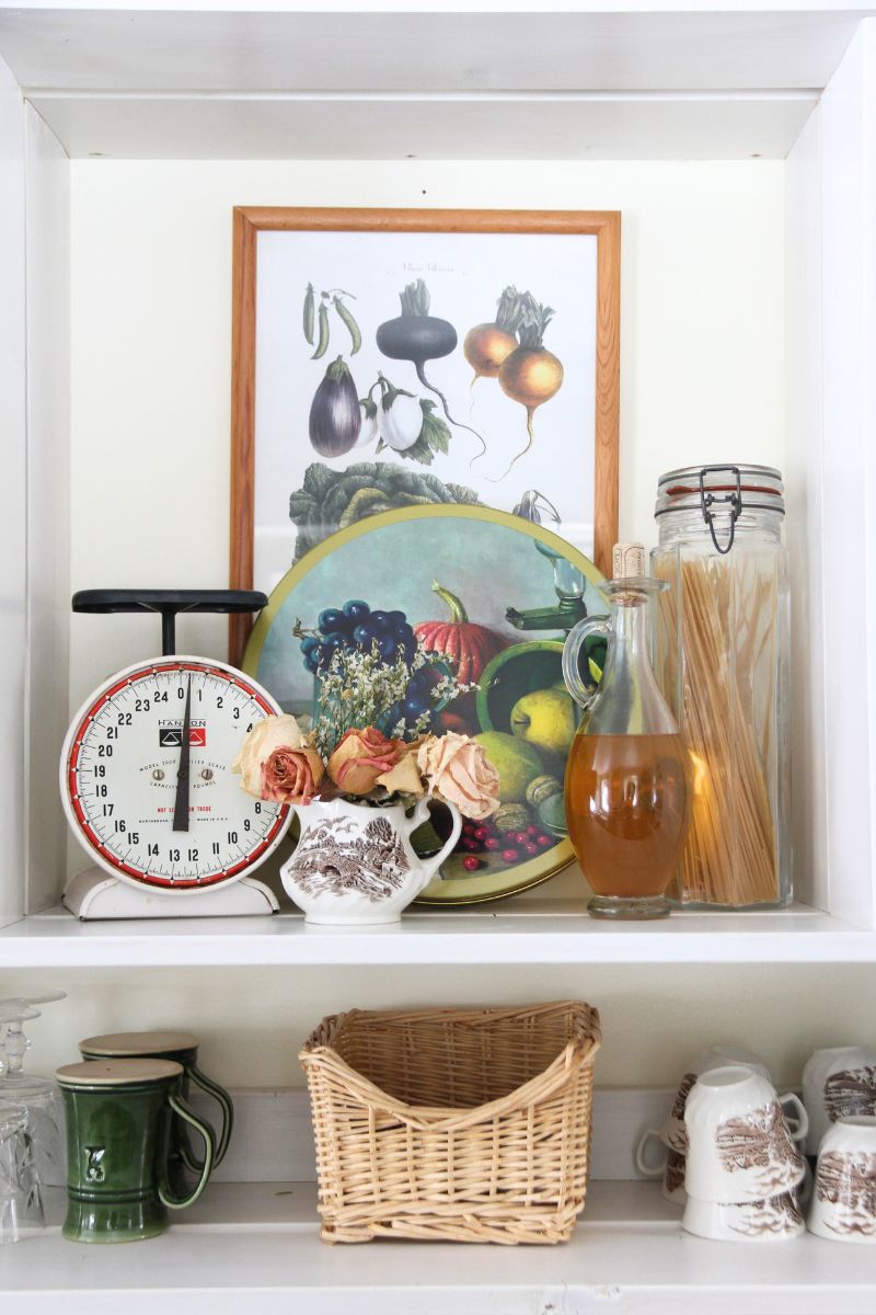 A decorative display on an open kitchen shelf, with a scales, round tin, spaghetti jar, a glass decanter of apple cider vinegar and a creamer pitcher filled with dried roses. 
