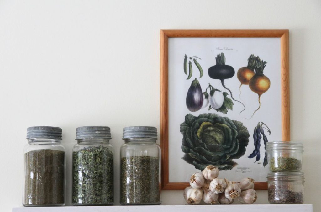 Vintage glass jars filled with dried herbs on a kitchen wall shelf with vegetable art in the background