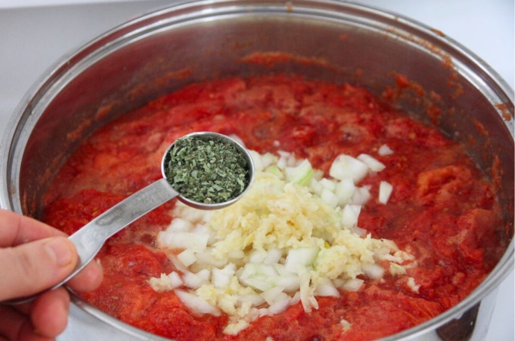 A pot of cooked tomatoes, with onions and herbs being added into the pot.