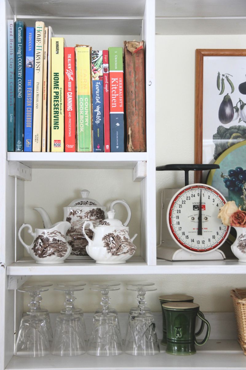 A large display shelf filled with cookbooks, a brown and white teapot, sugar and creamer, plus an old, white kitchen scale. 