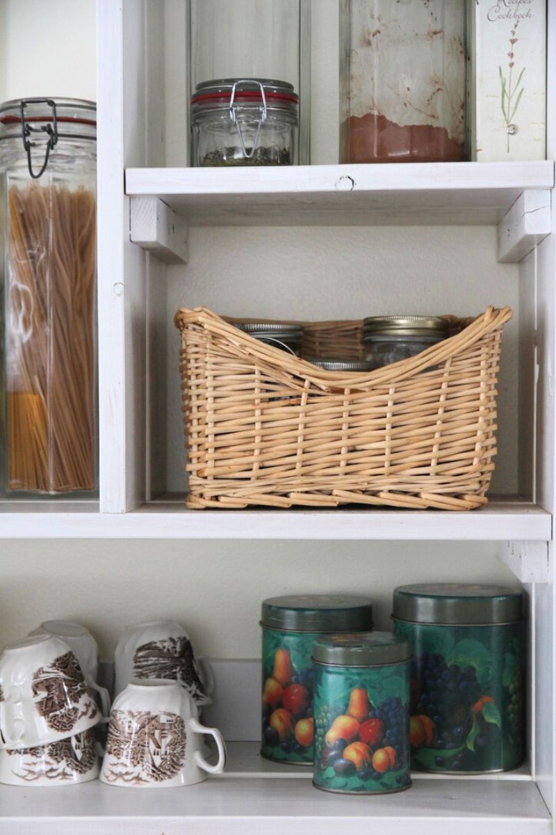 A small square basket holding small spice jars and containers on a white open kitchen shelf.