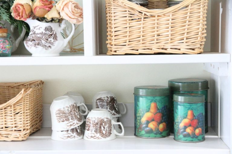 An open kitchen shelf with a trio of matching green food tins, brown and white china tea cups and woven wicker baskets