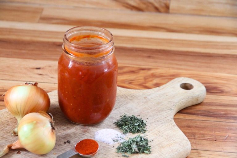 A jar of finished tomato sauce, sitting on a wooden cutting board with onions, salt and spices beside.