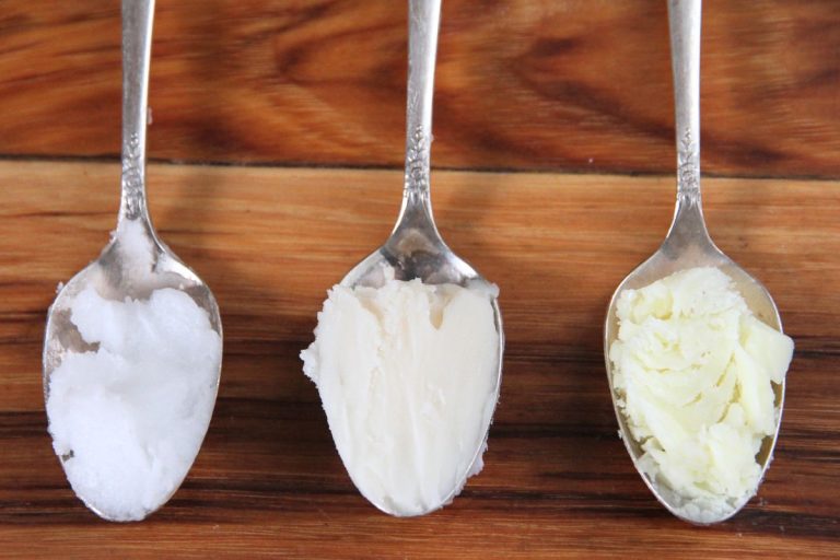 Three silver spoons holding coconut oil, lard and tallow by a cast iron pan.