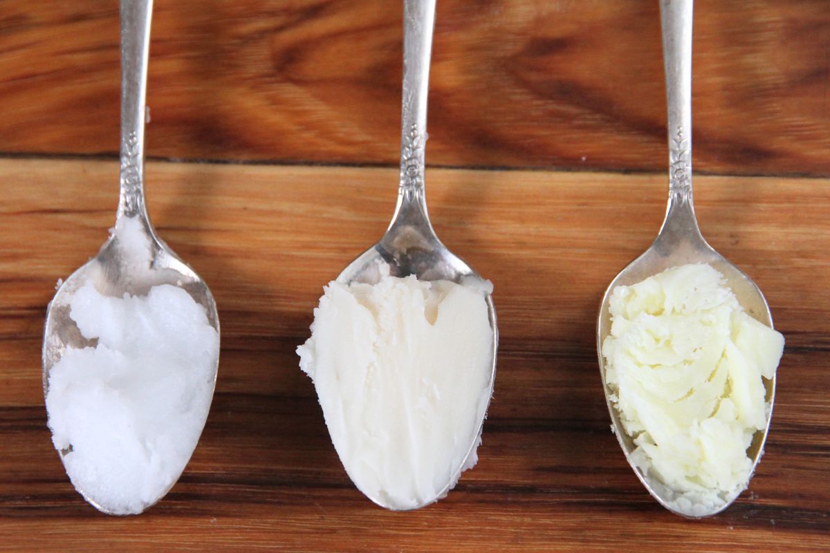 Three silver spoons holding coconut oil, lard and tallow by a cast iron pan.