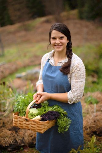 A woman standing in her garden in a blue apron with a square basket of vegetables
