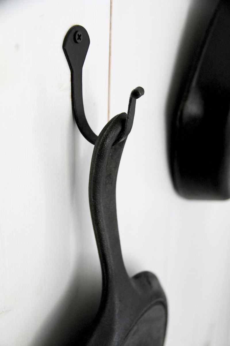 Pieces of cast iron hanging from a black hook in a whitewashed wood panel kitchen wall.