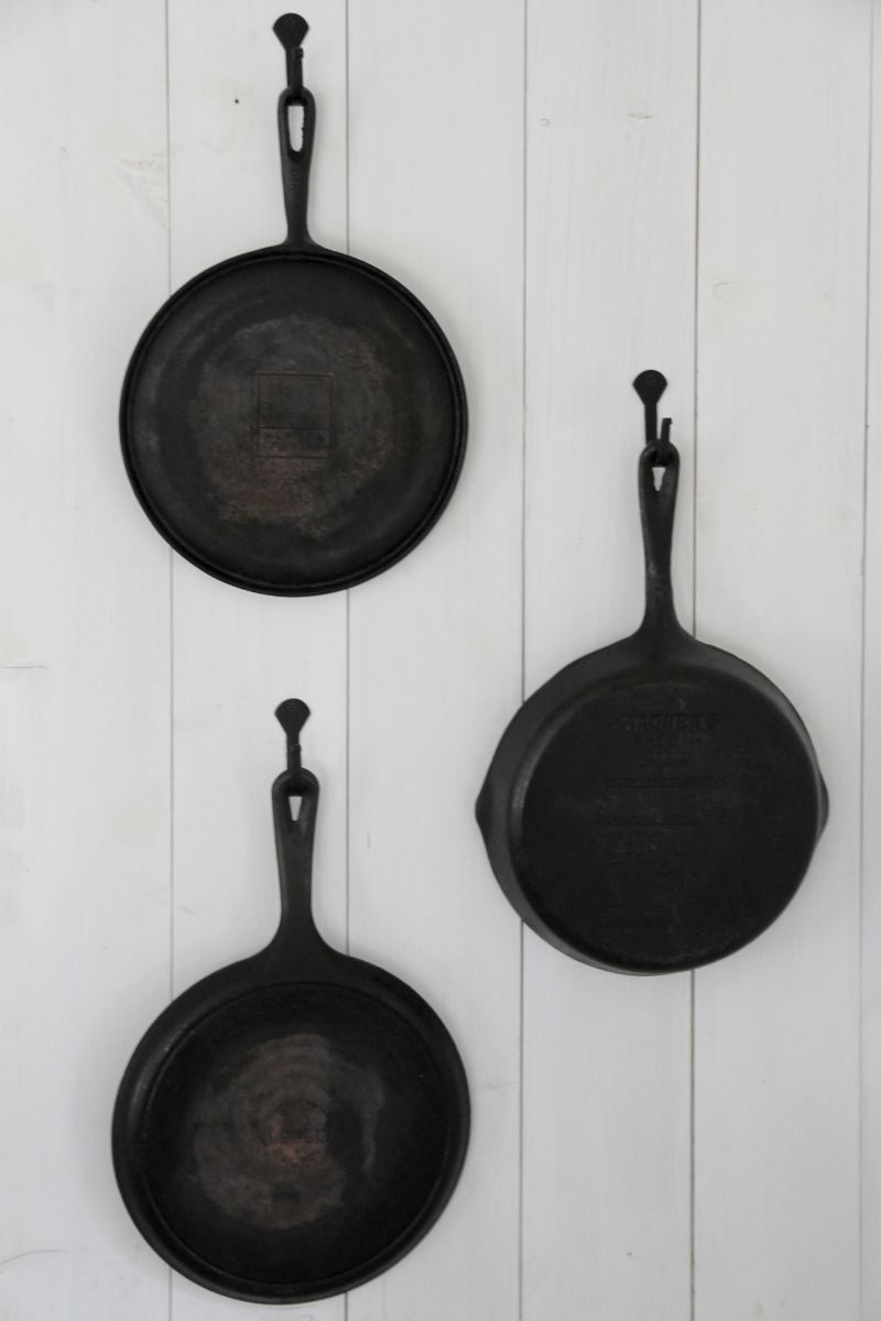 Three black cast iron pans hanging from hooks on a whitewashed wood panel wall.
