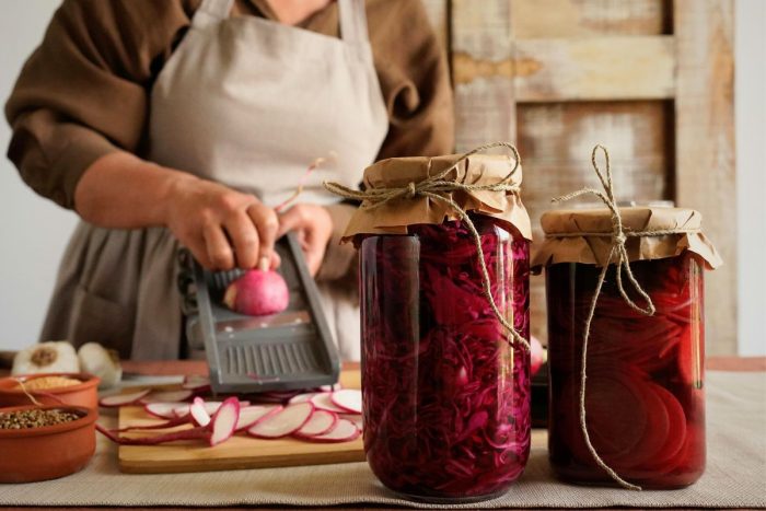 A woman in a dress and apron grating vegetables with jars of preserved vegetables on the side of the counter.