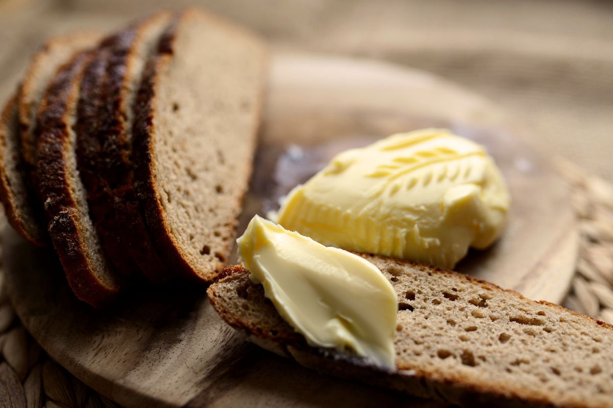 A loaf of brown bread being sliced on a wood cutting board with a side of butter.