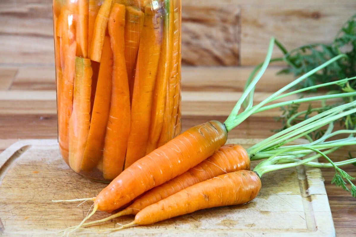 A jar filled with fermenting carrot sticks and a pile of fresh carrots in front of the jar.