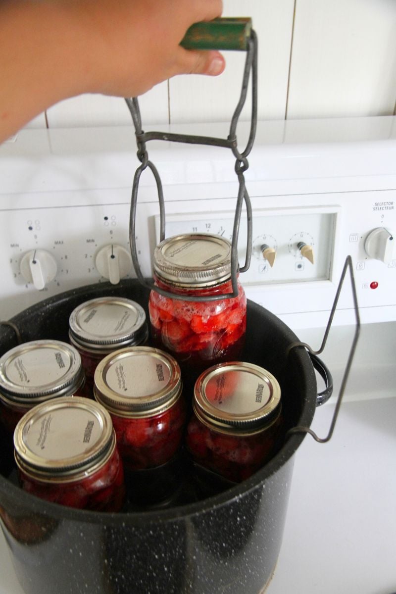 Lifting a jar of canned strawberry sauce from a small, boiling water pot. 