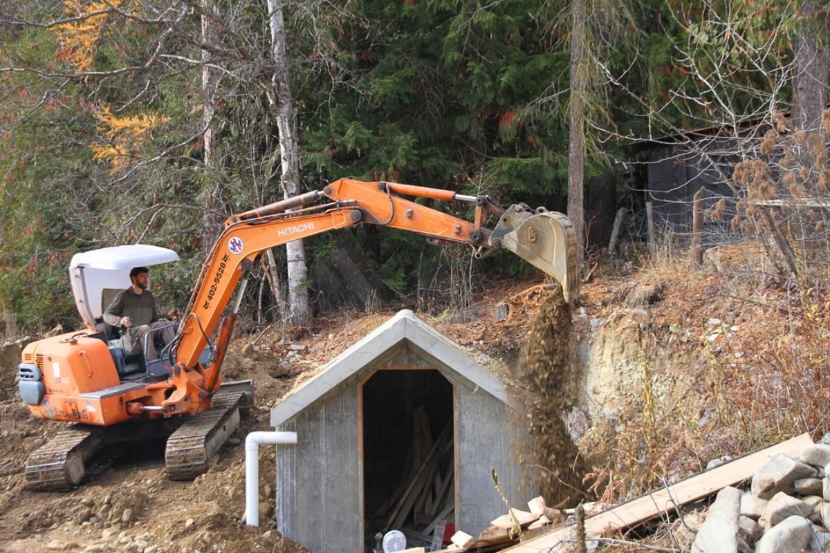 A builder, using a excavator to bury our concrete root cellar in the hillside where it was built. 