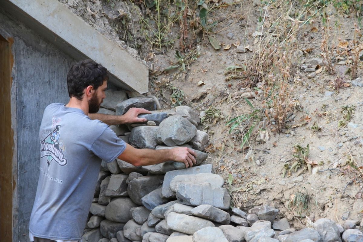 A man stacking round rock as a retaining wall on each side of the root cellar door. 