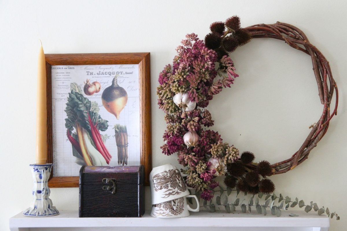 A herb wreath, stacked teacups, a candle and wooden box on a white kitchen shelf. 