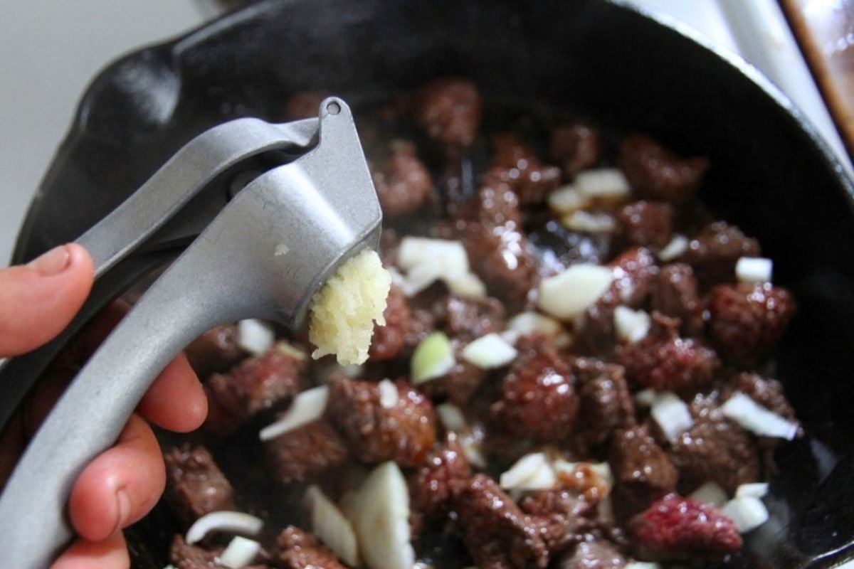 A garlic press, squishing garlic above the meat searing in a cast iron skillet.
