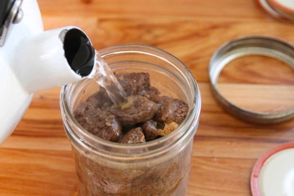 Pouring hot water from a white teakettle into a filled jar of seared bear meat.